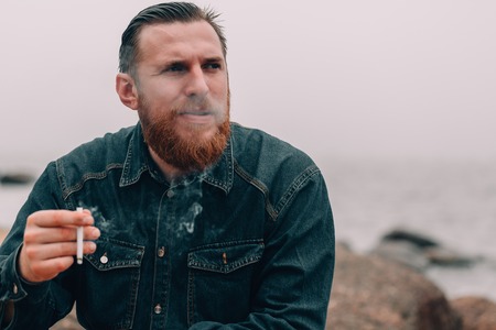 Serious bearded man smoking a cigarette on the beach. bearded hipster on the beach. Close-up portraitの写真素材