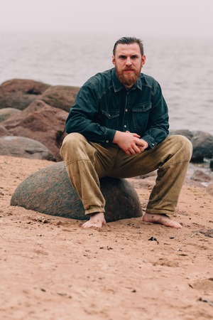 bearded man sitting on a rock near the beach. bearded hipster on the beachの写真素材