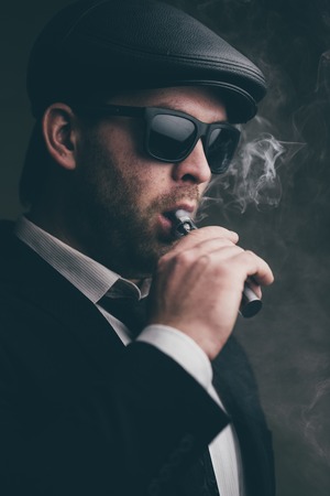Fashionable man in sunglasses and a leather cap smokes an electronic cigaretteの写真素材