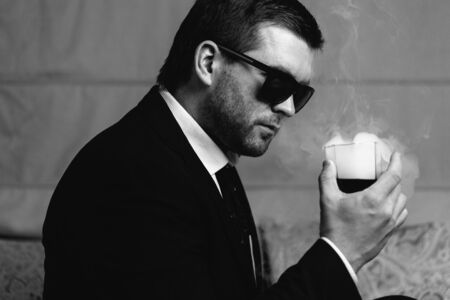 Stylish young man in sunglasses looking at steaming drink a glass in his handの写真素材