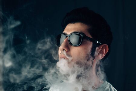 Young cool guy in sunglasses exhales a cloud of smoke. Studio horizontal portrait in close-up