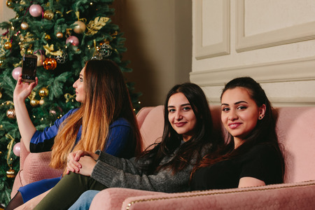 Three beautiful young girlfriends make selfie on a sofa near the Christmas tree. Studio horizontal portrait.の写真素材