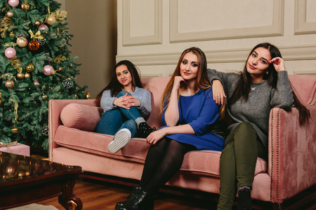 Three beautiful young girlfriends on a sofa near the Christmas tree. Studio horizontal portraitの写真素材