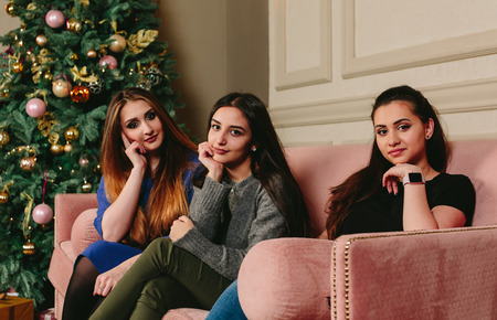 Three beautiful young girlfriends on a sofa near the Christmas tree. Studio horizontal portraitの写真素材
