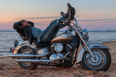 brutal man with a motorcycle on a sandy beach against the seaの写真素材