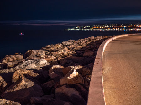 Night view of the promenade and the rocks along the illuminated french Mediterranean coast. At sea a fishing boat and in the background the lights of Mentonの写真素材