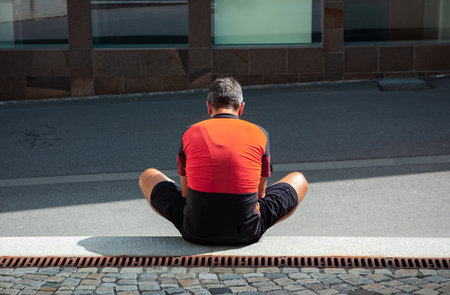 A lonely man in an orange-and-black cycling jersey sits on the floor in a sunny dayの写真素材