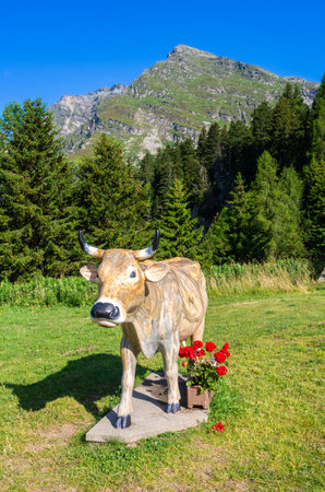 Maloja, Switzerland - July 10, 2023: Sculpture of a swiss cow with red flower at Maloja mountain pass in Switzerland. Beautiful green sunny morning landscapeのeditorial素材