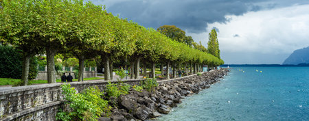 A scenic lakeside promenade with lush green trees and a stone walkway under a cloudy sky.