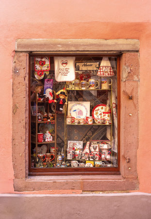Ribeauville, France - August 24, 2025: A quaint shop window display in Ribeauville featuring Alsace-themed souvenirs, including mugs, textiles, and figurines, set against a rustic stone frame.のeditorial素材