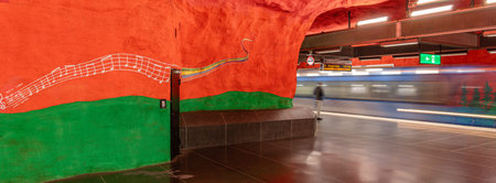 Stockholm, Sweden - June 9, 2025: Panoramic view of Solna centrum subway station in Stockholmwith a striking red rock wall, featuring colorful musical notes painted on it. A train is passing by.のeditorial素材