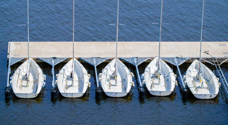 Aerial view of five white sailboats lined up at a dock on calm blue water.