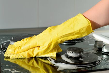 A hand in a yellow rubber glove washes a gas stove on a sunny day. Kitchen cleaning