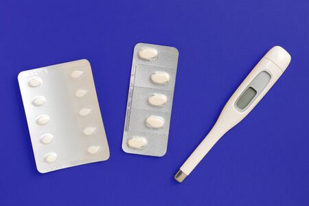 Electronic thermometer and pills on a deep blue background, top view. Healthcare concept