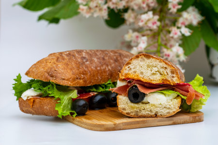 Sandwich from baked bread, mozzarella cheese, salami, prosciutto, olives, sauce, salad leaves, on a wooden bottomの写真素材