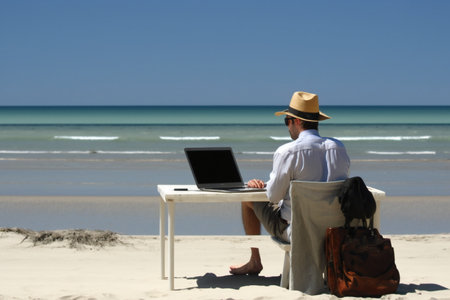 man working on a computer on the beach with a hat and bagの素材