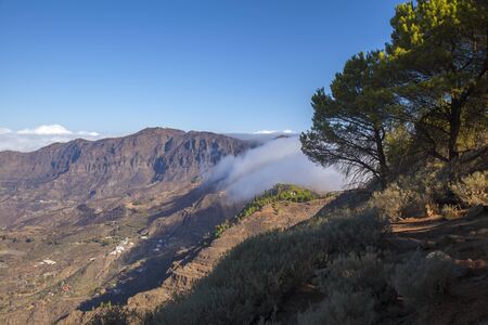 Gran Canaria, October, clouds flowing into Caldera de Tejeda from the right over mountain pass Cruz de Tejeda, Los Moriscos cliffs affected by wildfire of summer in the backgroundの写真素材