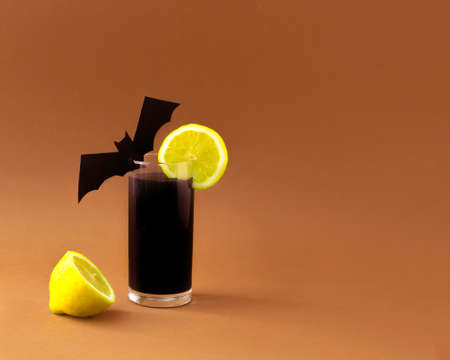 Black lemonade with bat and lemon on a glass. Halloween concept. On a brown autumn background.