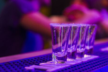 Tequila shots on the bar table at night party, people on the background, alcoholic full shots with...