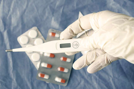 Body thermometer holding in hand, glove and doctors hand, drugs on the background