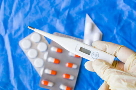 Body thermometer holding in hand, glove and doctors hand, drugs on the background