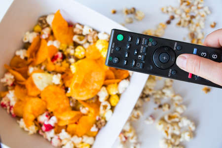 Fried popcorn and chips in bowl on the white background, hand holding TV controller, low angle viewの写真素材
