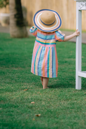 Back view of little girl in striped dress and straw hat standing on green grassの写真素材