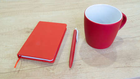 Red notepad and pen and ceramic mug cup for tea and coffe. On wooden desk.の写真素材