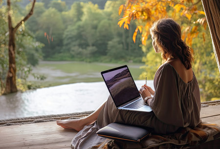 Young woman working on laptop sitting on the wooden floor by the riverの素材