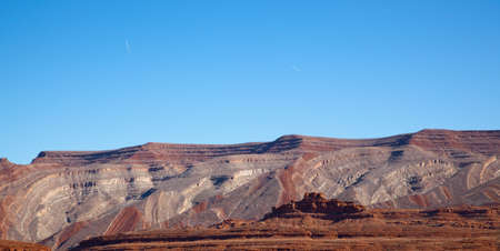 A natural mountain range seems to be sculpted by some larger force, showing color variations in the eroding rocks with a smaller sandstone formation in front lit by the morning sun and bright blue sky.の写真素材