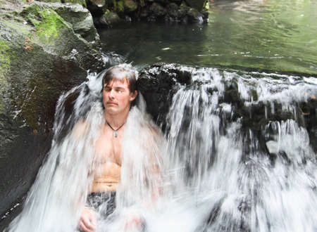 A young man sits at the base of a small waterfall relaxing as the hot thermal waters from Arenal Volcano create a silky flow over his body の写真素材