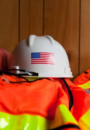A white hard hat with the American Flag is setting on top of a orange and reflective yellow safety vest and glasses in a construction trailer.のeditorial素材