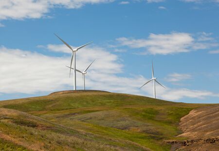 Looking up at three large windmills on top of a rolling green hill with blue sky and light clouds.