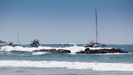 Boats are anchored in a cove of the Pacific Ocean on the rocky coast of Costa Rica with big waves...