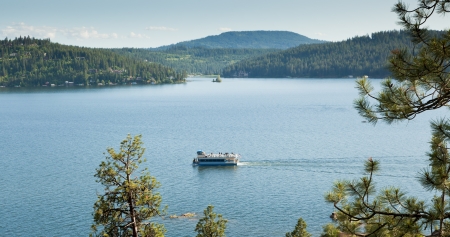 A tour boat cruises around Lake Coeur d'Alene, Idaho on blue water with background mountains and framed by pine trees.の写真素材