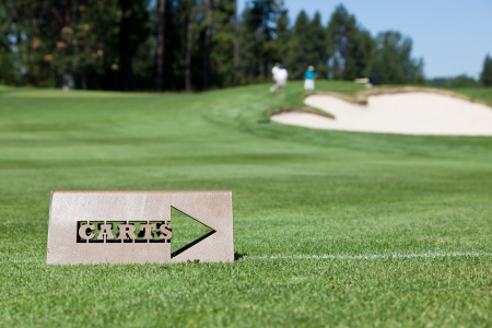 A cut out metal sign tells golf carts when to head back to the path off the green. Background of golfers by the sand trap and hole.の写真素材
