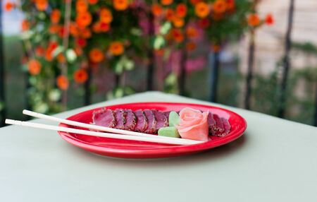 A rose made from pickled ginger with wasabi leaves sits on a red plate with seared tuna  Tabletop...