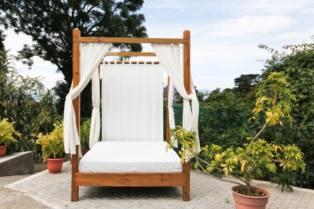 A wooden outdoor bed with white linens and privacy curtains on a brick patio at a tropical resort.の写真素材