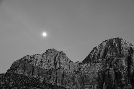A black and white image at twilight with a bright rising moon creating a soft glow above the mountains in Zion National Park, UT.の写真素材