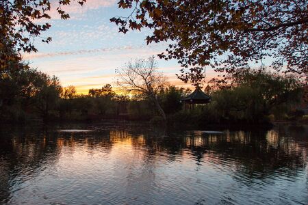 An orange glowing sunset over a park like setting of a calm pond with a bridge and gazebo and fall trees.の写真素材