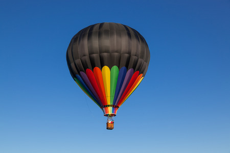 A black and rainbow hot air baloon floating on a calm clear blue sky day.の写真素材
