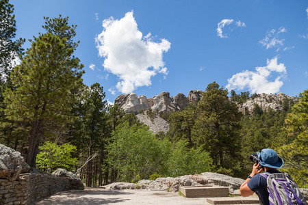 A young man stands at the base of the mountain to take a picture of Mount Rushmore with pine trees and blue sky.のeditorial素材