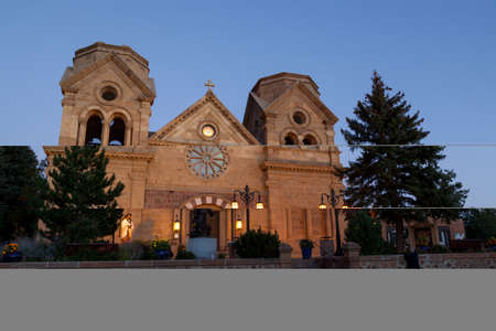 The Cathedral Basilica of St. Francis of Assisi located in Santa Fe, New Mexico, at twilight with soft glowing lights on the statues in the courtyard and vibrant blue sky.の写真素材