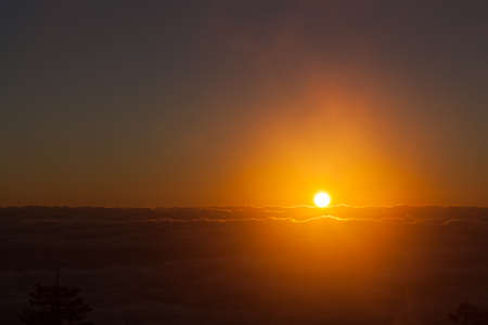 The sun looks like a glowing orange ball of fire as it lights up the fog and mist above a thick layer of clouds covering the valley by the Sandia Mountains in New Mexico.の写真素材
