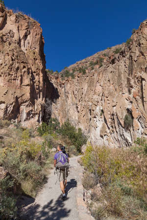 A man with a camera, tripod, and backpack walks on a pathway in Frijoles Canyon looking at the rock walls with cave dwellings in Bandelier National Monument, New Mexico.の写真素材