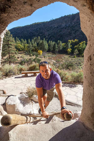 A smiling young man climbs up a wooden ladder leading to a cave dwelling entrance at the historical site of Bandelier National Monument in New Mexico.の写真素材