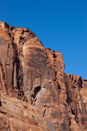 The towering red sandstone walls of Glen Canyon with several cracks through the aged rock lit by bright sunshine and topped with brilliant blue sky.の写真素材