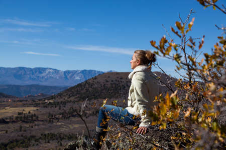 A woman enjoying the fall sunshine high up on a rock overlooking a valley with distant mountains in Utah near Zion National Park.の写真素材
