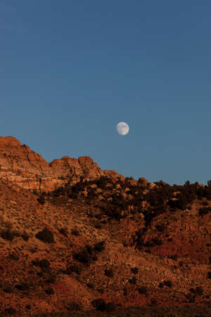 An almost full moon rising above red sandstone mountains with clear a blue evening sky near Zion National Park, Utah.の写真素材