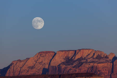 The full moon rising over the dramatic mountains of Zion National Park lit by the setting sun with a clear sky.の写真素材